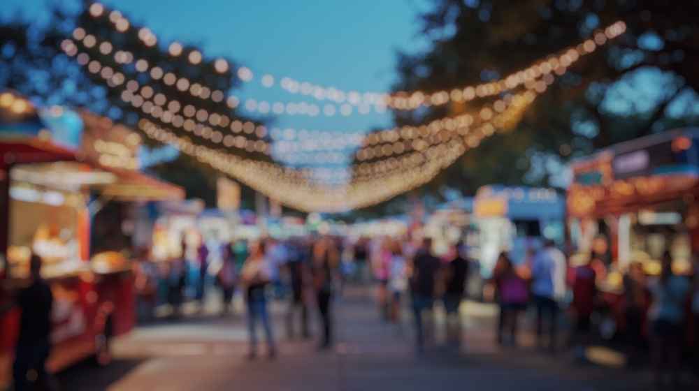 The image shows vibrant food vendors at an outdoor festival, with people enjoying the pleasant spring weather, surrounded by colorful stalls featuring environmentally friendly food packaging. Attendees are happily sampling various foods and beverages, contributing to a lively atmosphere that promotes positive change and sustainable practices.